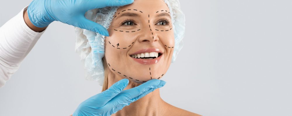 Cheerful excited middle aged woman in medical hat with pre surgery marks on her face looking at copy space for ad and smiling, surgeon hands in blue gloves touching female skin